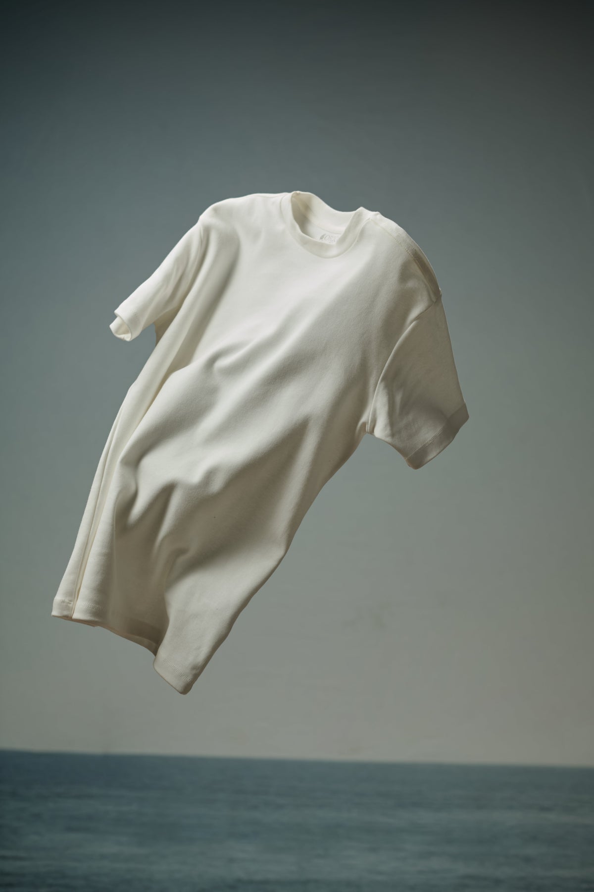 Camiseta masculina off-white de algodão suspensa no ar sobre o mar, em editorial ORIGGO